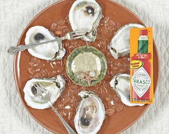 May include: A terracotta plate with six oysters on crushed ice, each with a small fork. A small green dish with seashell details sits in the center. A bottle of Tabasco brand pepper sauce is on the right.
