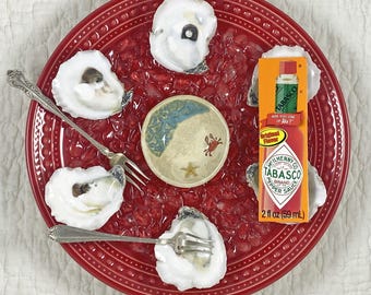 May include: A red plate with oysters, a small bowl with a beach scene, and a bottle of Tabasco sauce. Two forks are placed on the plate. The Tabasco bottle reads "TABASCO BRAND PEPPER SAUCE" and "2 fl oz (59 mL)".