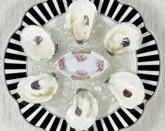 May include: A round plate with a black and white striped border holds five open oyster shells arranged in a circle around a small, oval, white porcelain dish decorated with pink floral patterns and gold trim, all resting on clear ice.