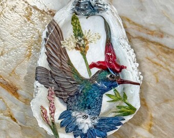 May include: A decorative oyster shell featuring a detailed painting of a hummingbird in flight with blue, green, and brown feathers. The bird is surrounded by pressed botanical elements, including a red flower, green leaves, and small white blooms.
