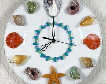 May include: White round clock with a variety of seashells and a starfish. The clock face features a ring of blue and green glass pieces. The clock hands are black.