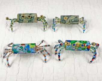 May include: Four decorative crab figurines crafted from cylindrical bodies with patterned paper surfaces and multi-colored beaded legs. Each crab features small circular eyes and articulated limbs made of various glass beads in shades of blue, green, yellow, and orange.