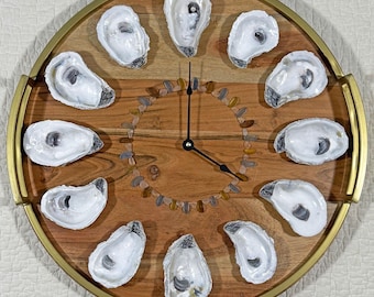 May include: A round wooden clock with a gold-colored rim and handles. The clock face is made of wood and features oyster shells marking the hours. Small stones form a circle around the clock's center. The clock hands are black.