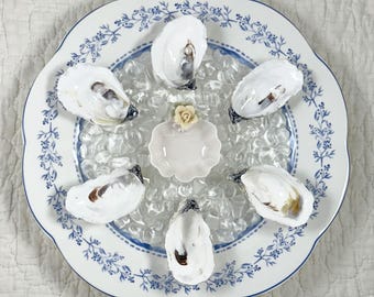 May include: A white and blue floral patterned plate with six oysters arranged around a small, pink, scalloped dish. The oysters are on a bed of clear ice. The plate has a scalloped edge.