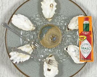 May include: A gray speckled plate with oysters, a small ceramic dish with a turtle design, and a bottle of Tabasco sauce. The oysters are arranged around the dish on a bed of ice, with two forks.
