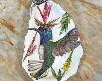 May include: Hand-painted oyster shell art featuring a hummingbird. The bird has blue, green, and brown feathers, with a long black beak. The shell is decorated with red and yellow flowers and green leaves.