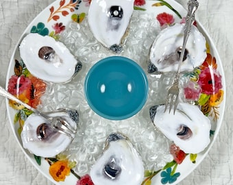May include: A round, floral-decorated serving plate with six oysters on ice, a small teal bowl, and two oyster forks. The oysters are open, revealing the oyster meat. The plate has a colorful floral pattern with red, orange, and blue flowers.