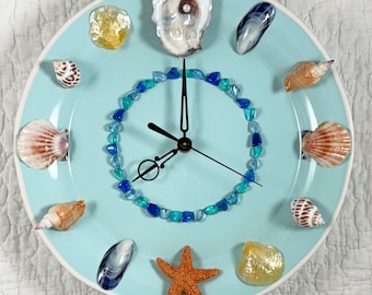 May include: A decorative wall clock with a light blue face, adorned with seashells, sea glass, and a starfish. The clock hands are black, and the numbers are represented by various seashells and sea glass pieces.