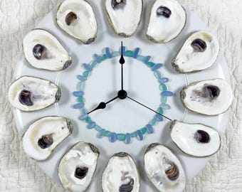 May include: A unique clock made from a white plate, featuring oyster shells as hour markers. The clock face has a blue and turquoise bead design around the center, with black clock hands. The shells have a natural, off-white color.