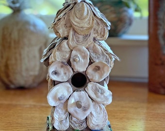 May include: A birdhouse covered in layered, natural oyster shells with a central circular entrance hole. The base is decorated with small, smooth, multi-colored sea glass pieces in shades of green, blue, and brown, arranged in a single row.