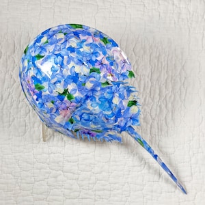 May include: A decorative, floral-patterned object resembling a horseshoe crab. The object is covered in blue, purple, and white hydrangea flower designs. The tail is long and pointed. The object is displayed on a white, quilted surface.