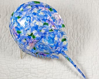 May include: A decorative, floral-patterned object resembling a horseshoe crab. The object is covered in blue, purple, and white hydrangea flower designs. The tail is long and pointed. The object is displayed on a white, quilted surface.