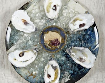 May include: A ceramic plate with a blue and white glaze, holding six oyster shells and a small decorative bowl. The oysters are arranged around the bowl, which features a floral design. Clear glass beads fill the plate.