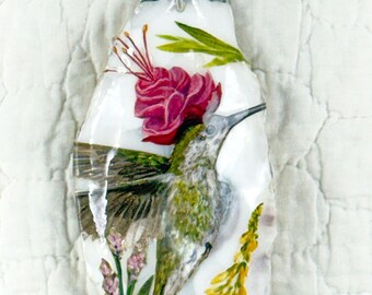 May include: A decorative oyster shell pendant featuring a hand-painted hummingbird in shades of green and brown, hovering near a vibrant pink flower. The shell is accented with small yellow and purple floral details and hangs from a beaded cord.