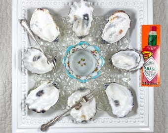 May include: A square white serving platter with a beaded edge holds seven oyster shells arranged around a central floral-patterned porcelain bowl. Two silver oyster forks are placed on the platter, alongside a 2 fl oz bottle of Tabasco sauce.