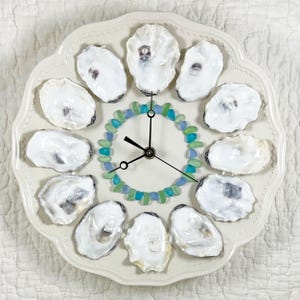 May include: A decorative clock featuring a white ceramic plate with a scalloped edge. The clock face is adorned with twelve oyster shells, blue and green glass beads, and black clock hands. The clock is a unique coastal home decor item.