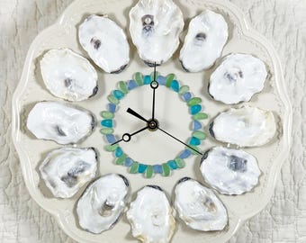 May include: A decorative clock featuring a white ceramic plate with a scalloped edge. The clock face is adorned with twelve oyster shells, blue and green glass beads, and black clock hands. The clock is a unique coastal home decor item.