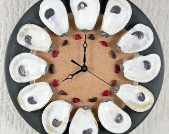 May include: A decorative wall clock featuring a dark gray circular frame. The clock face is a light brown color, with oyster shells marking the hours. Red and dark red stones are scattered around the clock face. The clock hands are black.