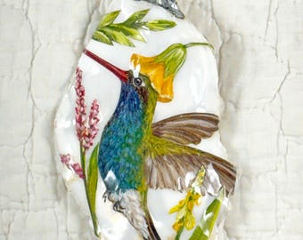 May include: Hand-painted hummingbird pendant on a white, irregular-shaped shell. The bird has blue, green, and brown feathers, a red beak, and is surrounded by yellow flowers and pink blossoms. A silver-colored bail is attached to the top.