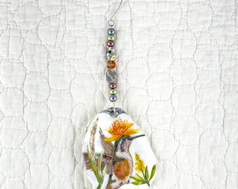 May include: A decorative hanging ornament featuring a painted oyster shell with a hummingbird and yellow flower illustration. The shell is suspended by a silver wire hook adorned with a vertical strand of multicolored glass and metallic beads.