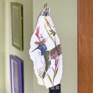 May include: A decorative hanging ornament crafted from a white oyster shell featuring a detailed illustration of a hummingbird with blue, green, and brown plumage, surrounded by painted pink and yellow botanical elements and a small metal ring.