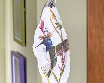May include: A decorative oyster shell ornament featuring a hand-painted hummingbird with a blue head, green body, and brown wings. The bird is surrounded by green stems and pink floral accents. A small metal ring is attached to the top for hanging.