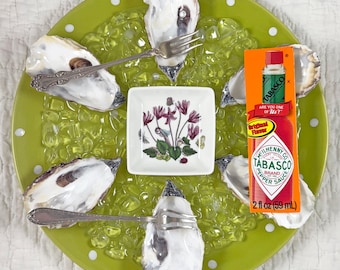 May include: A lime green plate with white polka dots holds six oyster shells, each with a fork. A small square dish with a floral design sits in the center. A bottle of Tabasco sauce is on the plate.