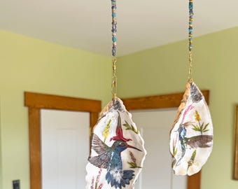 May include: Two hanging ornaments made from natural oyster shells, each featuring a detailed illustration of a hummingbird among flowers. The shells are suspended by vertical strands of blue, silver, and gold beads with metal hardware.