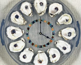 May include: A circular gray wooden tray with two side handles functioning as a wall clock. Twelve white oyster shells are arranged in a circle around a center ring of multicolored polished stones. Black clock hands are mounted in the middle.