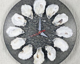 May include: A round, dark gray clock with a unique design. The clock face features a bed of small, dark gray stones and is encircled by twelve white oyster shells. The clock hands are silver with a red second hand.