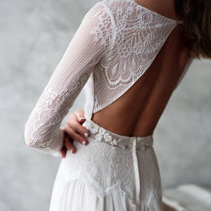 May include: White lace wedding dress with long sleeves and a low back. The dress features intricate floral lace detailing on the sleeves, bodice, and waist. The skirt is made of a flowing, lightweight fabric.