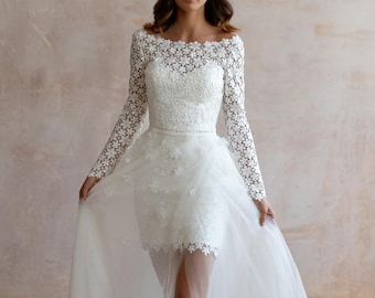 May include: A white wedding dress with a floral lace bodice and long sleeves. The dress features a short skirt with a sheer overlay and a long, flowing train. The bride is wearing nude heels.