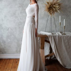 May include: A white wedding dress with a long, flowing skirt and long lace sleeves. The dress features a fitted bodice with a cutout detail at the waist. The bride is standing in a room with a wooden floor and a neutral-toned wall.