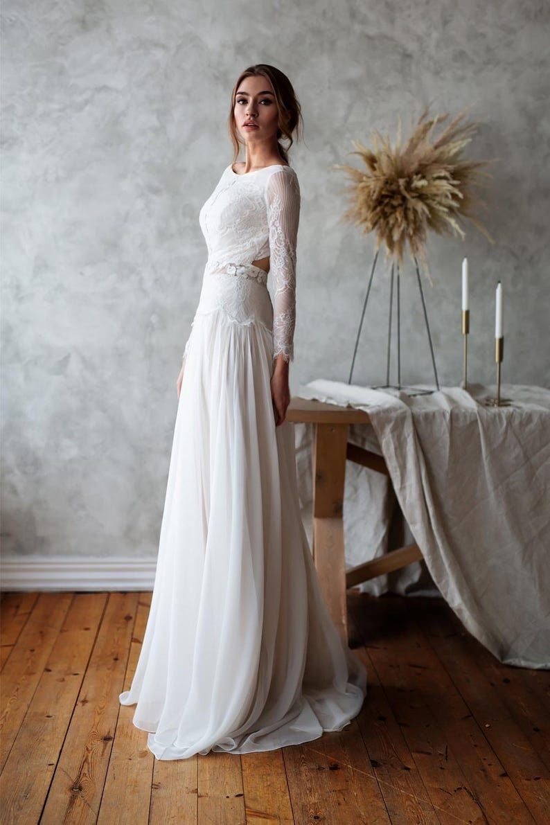 May include: A white wedding dress with a long, flowing skirt and long lace sleeves. The dress features a fitted bodice with a cutout detail at the waist. The bride is standing in a room with a wooden floor and a neutral-toned wall.