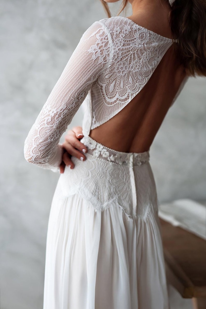 May include: White lace wedding dress with long sleeves and a low back. The dress features intricate floral lace detailing on the sleeves, bodice, and waist. The skirt is made of a flowing, lightweight fabric.
