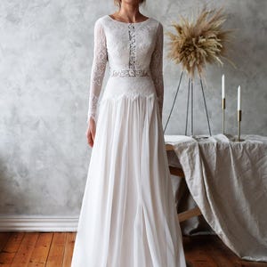 May include: A white wedding dress with long lace sleeves and a flowing skirt. The dress features a detailed lace bodice and a delicate belt. The bride is standing in front of a neutral background with wooden floors.