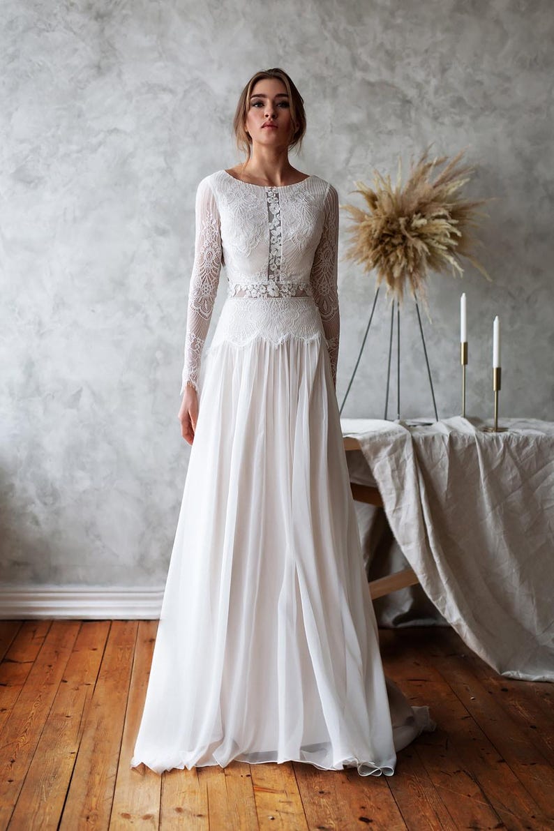 May include: A white wedding dress with long lace sleeves and a flowing skirt. The dress features a detailed lace bodice and a delicate belt. The bride is standing in front of a neutral background with wooden floors.