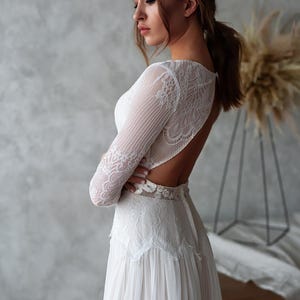 May include: A white wedding dress with long lace sleeves and an open back. The dress features a detailed lace bodice and a flowing skirt. The bride's arms are crossed, and her hair is pulled back in a ponytail.