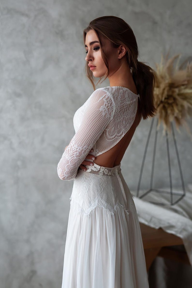 May include: A white wedding dress with long lace sleeves and an open back. The dress features a detailed lace bodice and a flowing skirt. The bride's arms are crossed, and her hair is pulled back in a ponytail.