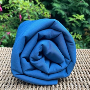 May include: Rolled-up bolt of solid teal fabric. The fabric is tightly wound, showcasing its smooth texture and deep blue-green color. The fabric is resting on a woven surface, with green foliage in the background.