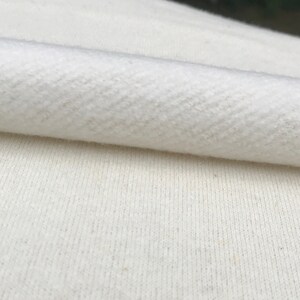 May include: Close-up of folded, off-white fabric with a textured herringbone weave. The fabric appears soft and possibly made of a natural fibre. The image highlights the texture and colour variations of the material.