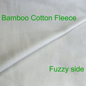 May include: Close-up of a white bamboo cotton fleece fabric. The fabric has a smooth side and a fuzzy side. The text "Bamboo Cotton Fleece" is printed on the smooth side and the text "Fuzzy side" is printed on the fuzzy side.