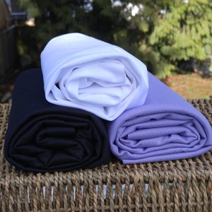 May include: Three rolls of fabric in different colors: white, black, and lavender. The fabric is folded and stacked on top of a woven basket.