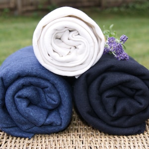 May include: Three rolled blankets in cream, navy blue, and dark navy blue are stacked on a woven surface. A sprig of purple flowers rests on top. The blankets are tightly rolled, showing their soft texture. The scene is set outdoors.