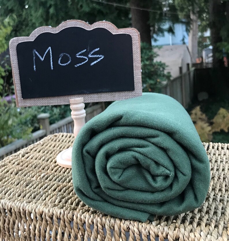 May include: A roll of dark green fabric labeled "Moss" on a woven wicker surface.