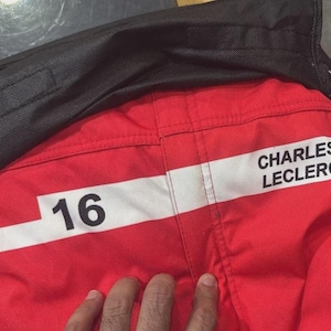 May include: Red racing suit detail with the number 16 and the name "CHARLES LECLERC" in white block letters. The suit has a white stripe and a black collar.