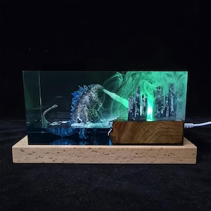 Könnte beinhalten: Eine Harzskulptur von Godzilla, der eine Stadt zerstört, mit einem grünen Leuchten, das vom atomaren Atem des Monsters und der Stadt ausgeht. Die Skulptur wird von unten mit einem blauen Licht beleuchtet und auf einem Holzsockel präsentiert.
