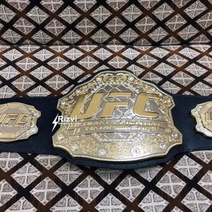 May include: A gold and silver UFC championship belt with black leather straps and the words "Ultimate Fighting Championship" on the front. The belt has two smaller UFC logos on the sides.