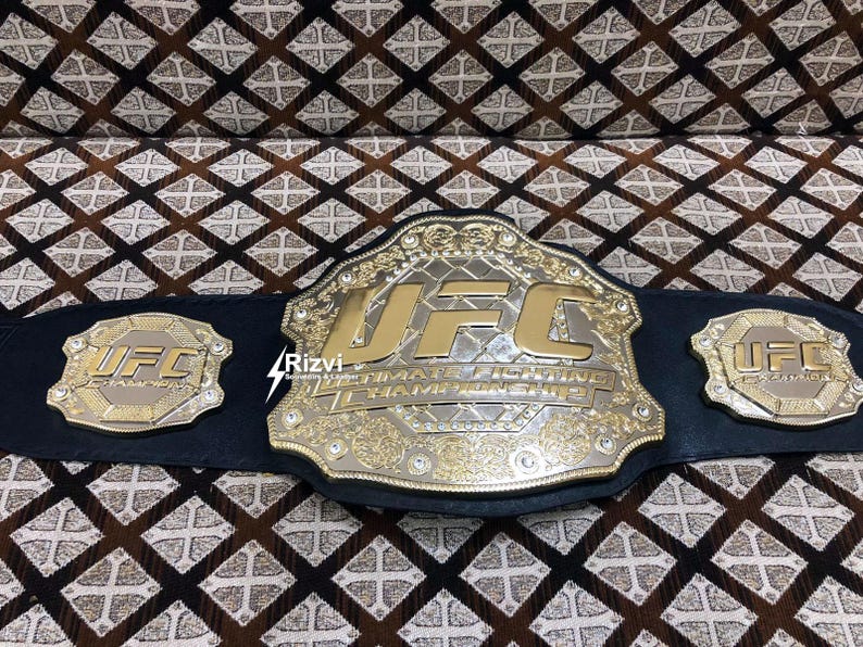 May include: A gold and silver UFC championship belt with black leather straps and the words "Ultimate Fighting Championship" on the front. The belt has two smaller UFC logos on the sides.