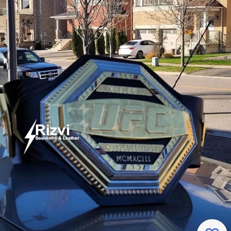 May include: A black and silver UFC championship belt with a gold octagon-shaped plate featuring the UFC logo and the text "MCMXCIII".
